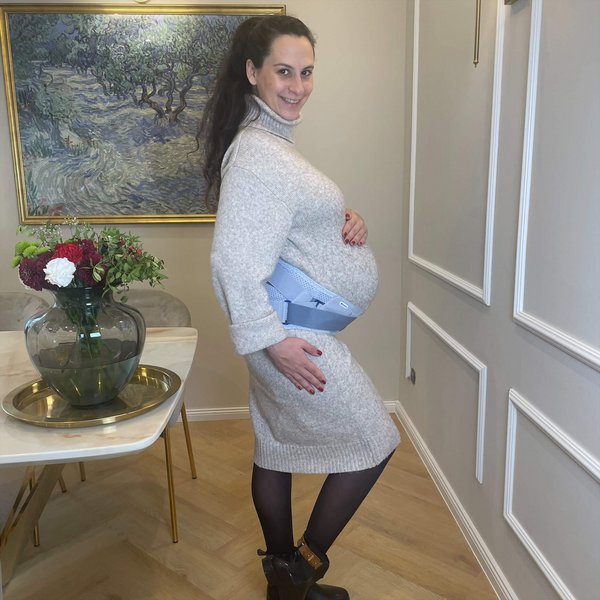 Zwangere vrouw staat in een elegant ingerichte ruimte, draagt een lichte gebreide jurk, zwarte panty, enkellaarsjes en een zwangerschapsorthese om haar buik.
