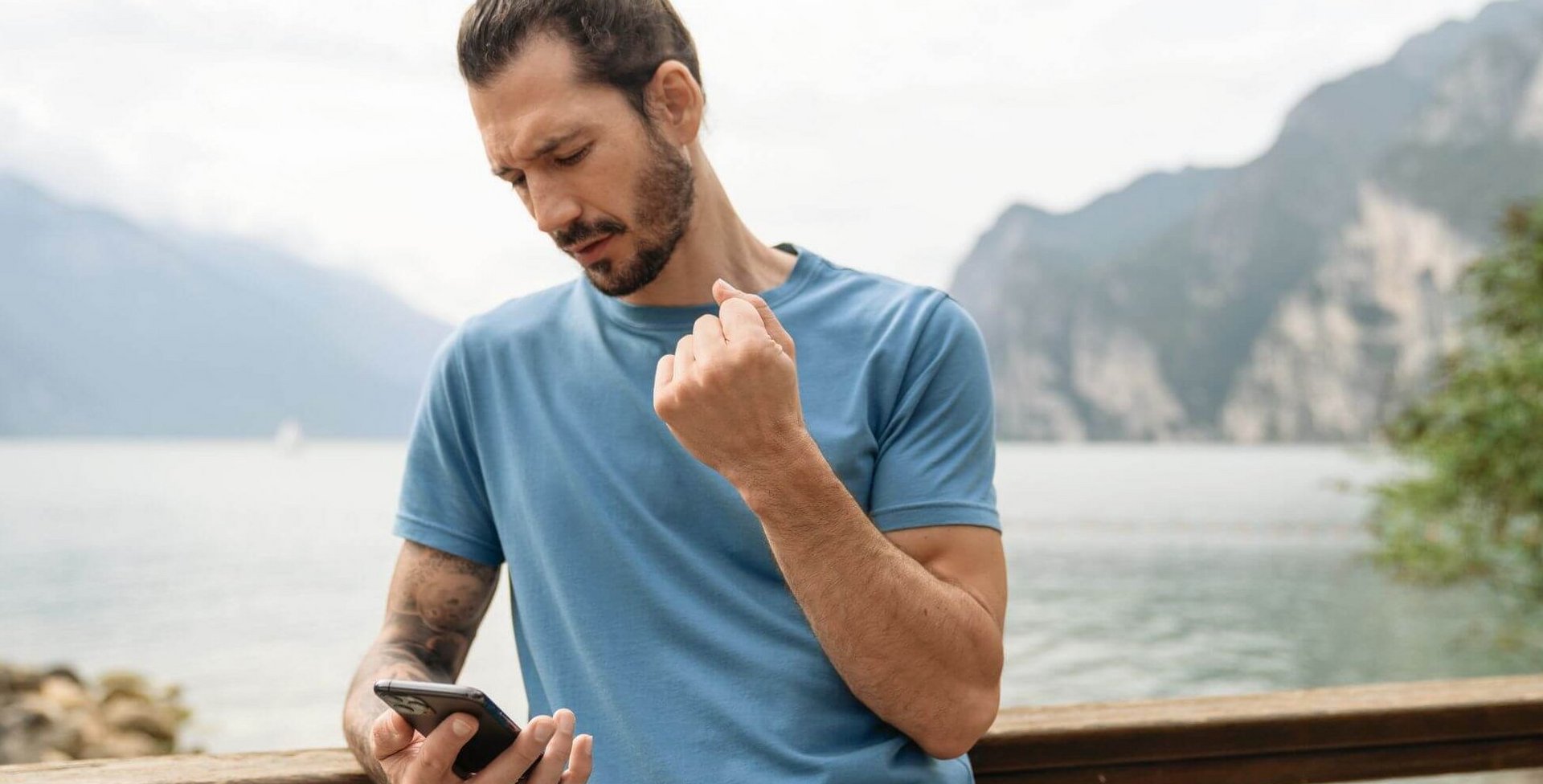 Op de foto zie je een man met smartphone die aan de oever van een bergmeer staat. Hij houdt zijn rechterarm met pijnlijke pols omhoog.