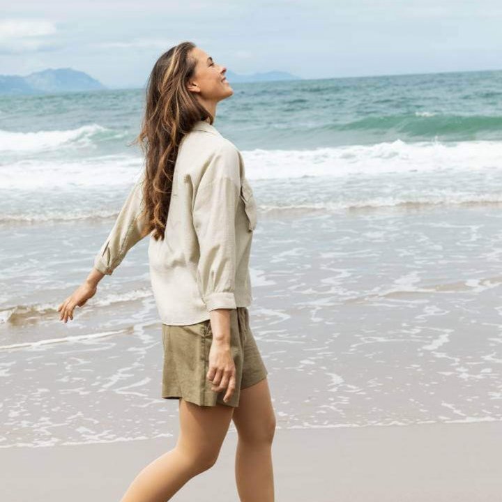 Een vrouw draagt VenoTrain soft compressiekousen en wandelt ontspannen langs het strand. Ze draagt een beige hemd, een korte olijfgroene broek en witte sneakers.