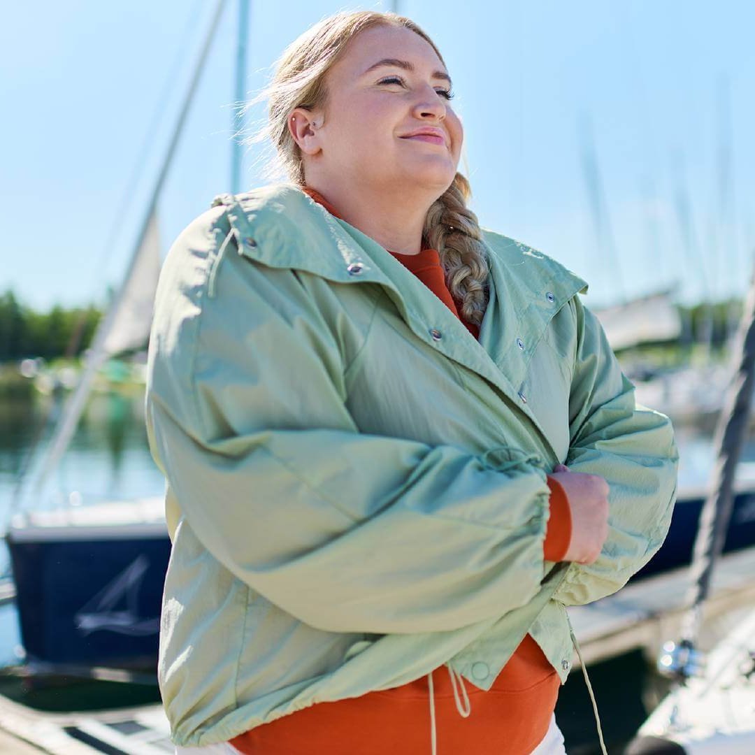 Vrouw met gevlochten haar staat ontspannen in een havenomgeving. Ze draagt een mintgroene jas over een oranje bovenstuk. Op de achtergrond zijn boten en masten te zien, die een rustige en maritieme sfeer scheppen.