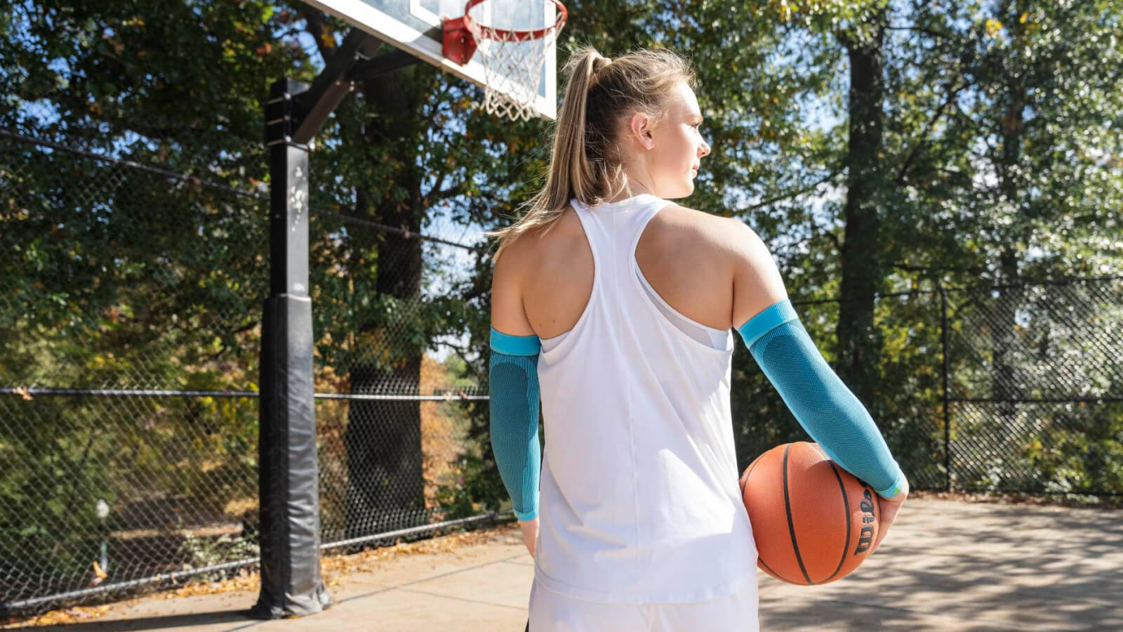Een vrouwelijke basketbalspeelster staat op een buitenveld met een basketbal in haar rechterhand. Ze draagt lichtblauwe compressie-armhoezen en kijkt geconcentreerd naar de basket.