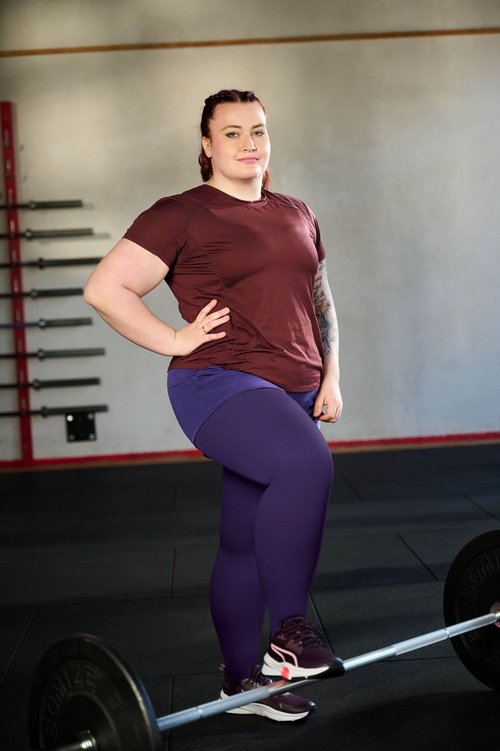 Sportief geklede vrouw met rood T-shirt en paarse legging staat zelfverzekerd in een sportschool met een been op een halterstang. Op de achtergrond zijn meerdere halterstangen aan een muurhouder zichtbaar.