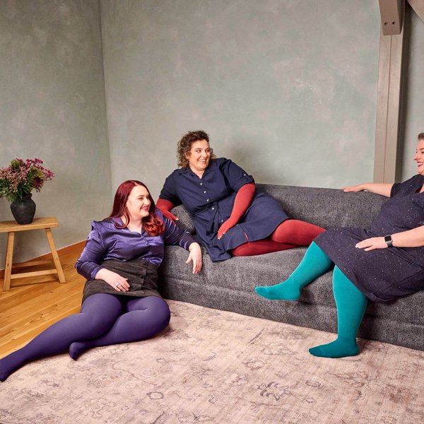 Drie vrouwen zitten ontspannen op een donkergrijze bank in een moderne woonkamer. Ze dragen kleurrijke panty's in paars, rood en turquoise bij donkere jurken of rokken en praten vrolijk met elkaar.