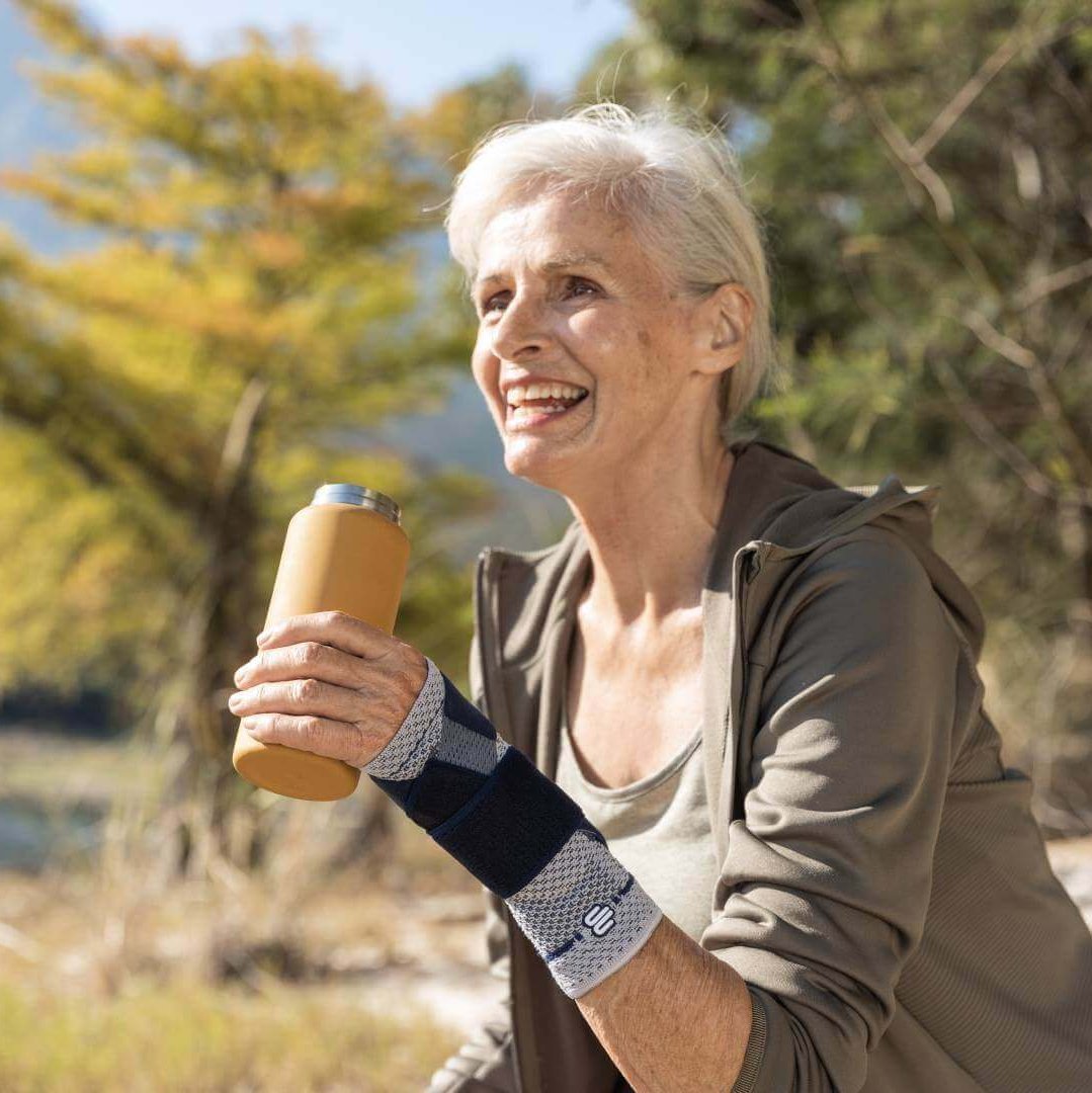 Op de foto is een oudere vrouw met een drankje bij een meer te zien, die tijdens het wandelen een ManuTrain polsbandage draagt om pijn in de pols te verlichten.