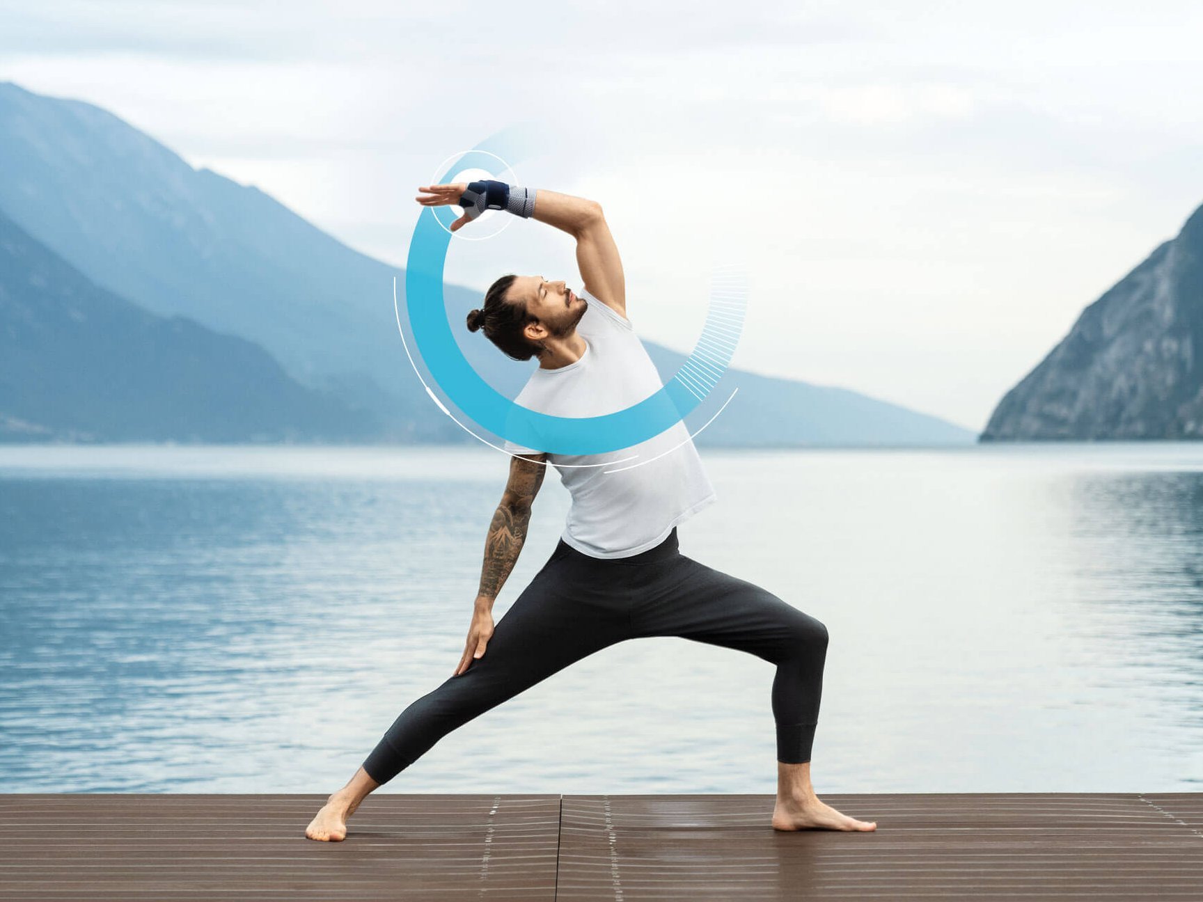 Persoon in sportkleding draagt de ManuTrain handbandage van Bauerfeind en doet een yogahouding op een houten steiger voor een bergmeer met een bewolkte hemel.