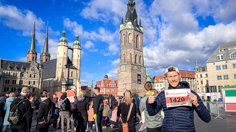 Een deelnemer van de Mitteldeutscher Halbmarathon houdt zijn startnummer omhoog op de marktplaats van Halle (Saale). Achter hem zijn de historische Rode Toren en Marktkirche te zien. Op de voorgrond bereiden lopers en toeschouwers zich voor op de race.