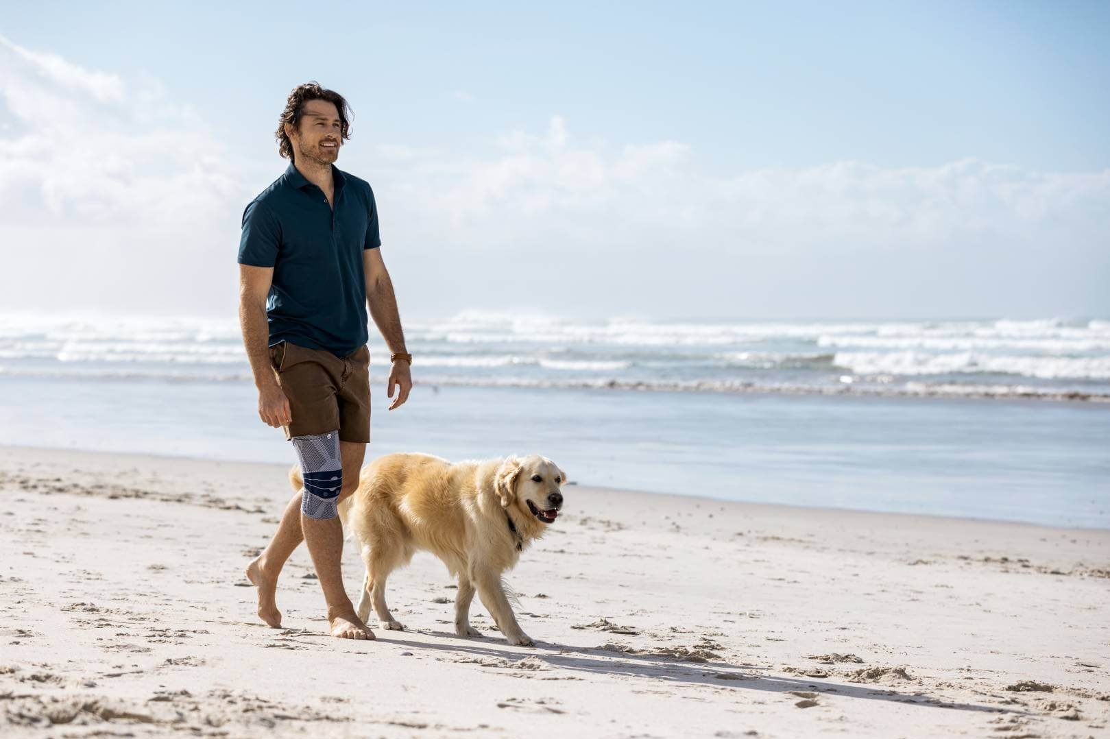 Een man wandelt blootsvoets met een golden retriever over een zonnig zandstrand. Hij draagt een GenuTrain kniebandage van Bauerfeind, terwijl op de achtergrond golven de oever bereiken.