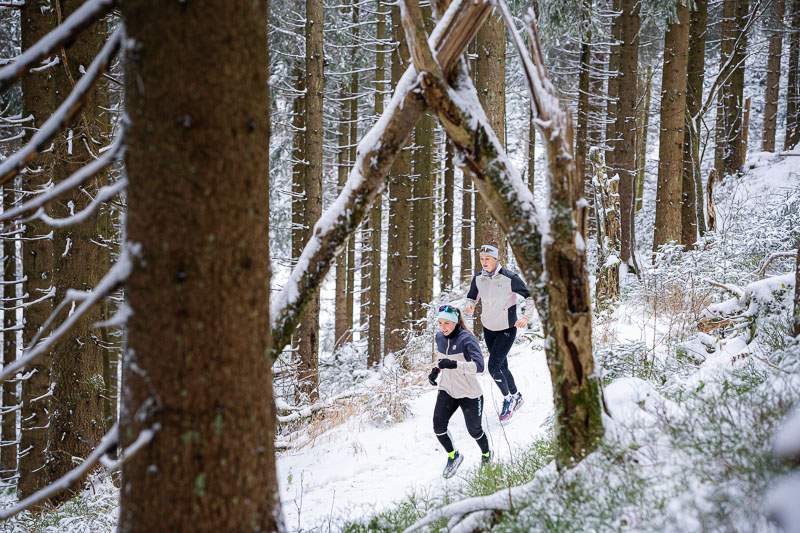 Twee hardlopers rennen door een besneeuwd bos. Het pad wordt omringd door hoge met sneeuw bedekte sparren, terwijl een natuurlijke boog van takken over het pad buigt. Beiden dragen warme hardloopkleding en hoofdbanden.