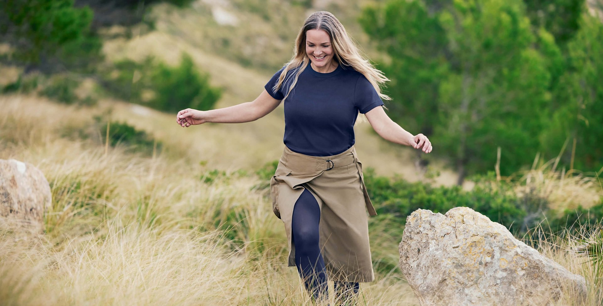 Een lachende vrouw met lang, los haar loopt lichtvoetig door een heuvelachtig natuurlandschap met hoog gras en rotsen. Ze draagt een nonchalant donkerblauw T-shirt, een kakikleurige wikkelrok en VenoTrain compressiekousen in donkerblauw.
