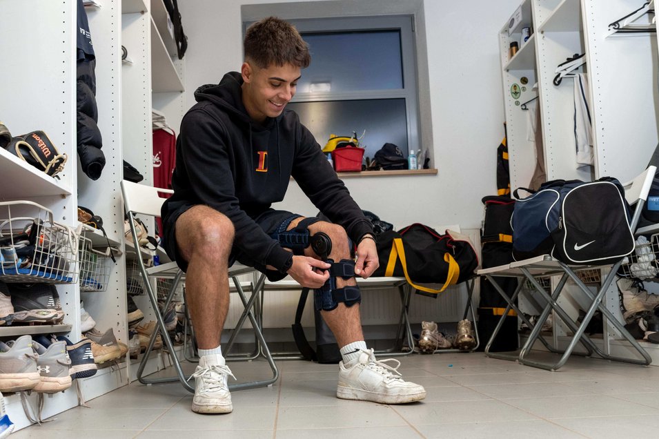 Simon Liedtke zit in de kleedkamer en doet de SecuTec Genu Flex knieorthese aan. Om hem heen zie je sporttassen, schoenen en honkbaluitrusting. Hij lacht geconcentreerd terwijl hij de orthese aanpast.