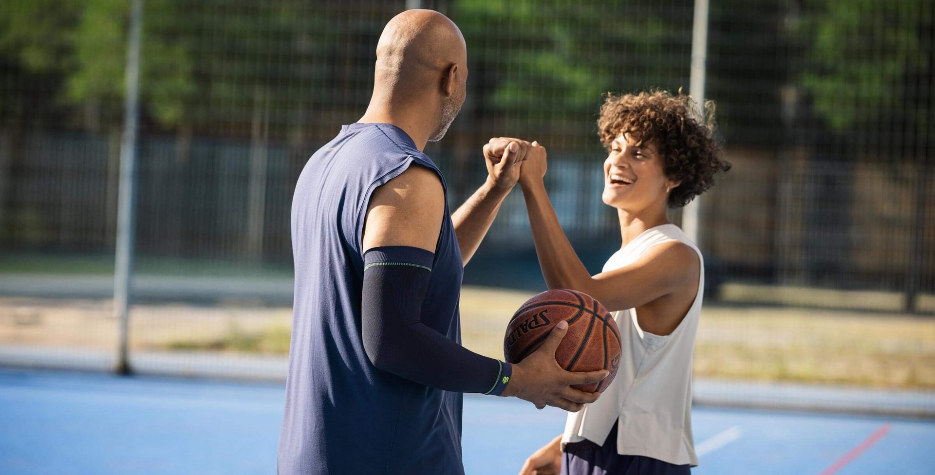 Twee basketballers op een blauw buitenveld geven elkaar lachend een boks. De persoon links, groter en met een kaal hoofd, houdt een basketbal vast en draagt een mouwloos shirt. De persoon rechts heeft krullend haar, draagt een wit sporttop en oogt vrolijk