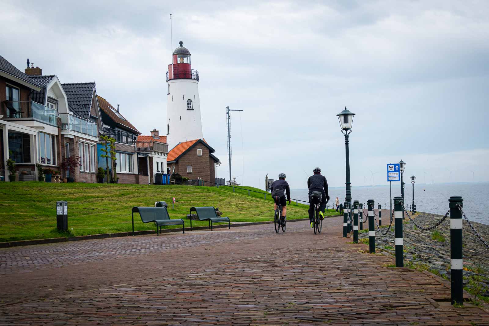 Twee ultrawielrenners van achteren die langs een promenade rijden waar een vuurtoren staat.