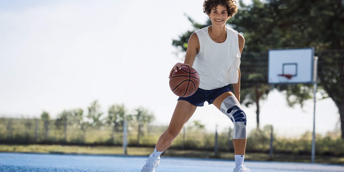 Jonge vrouw met wit hemdje, donkerblauwe sportbroek en witte kniebandage speelt basketbal op een buitenveld met blauwe ondergrond; ze dribbelt met de bal en lacht in de camera, op de achtergrond zijn een basketbalring en bomen te zien.