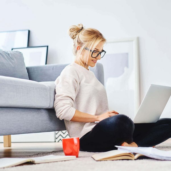 De afbeelding toont een jonge vrouw die met haar laptop op de grond van haar woning zit en werkt.