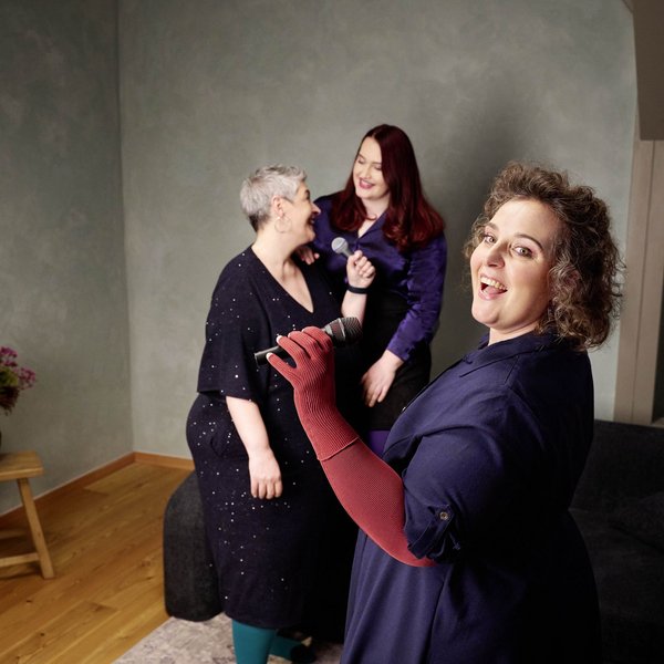 Drie vrouwen staan in een woonkamer met lichtgroene wand en zingen samen met microfoons. De vrouw op de voorgrond draagt een donkerblauwe jurk en rode compressiehandschoenen; achter haar lachen twee andere vrouwen.