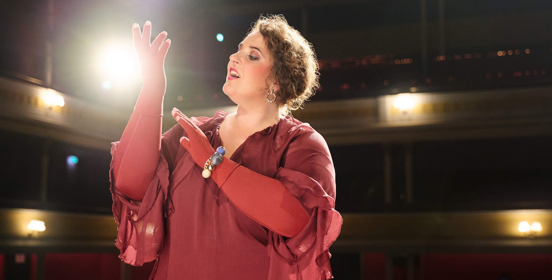 Close-up van een vrouw in een bordeauxrode jurk met bijpassende handschoenen, die met een expressief gebaar haar handen heft terwijl ze zingt. Op de achtergrond zie je vage lichten van de theaterzaal.
