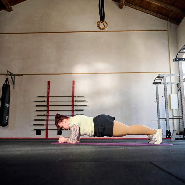 Persoon doet een onderarmsteun-oefening (plank) op een fitnessmat in een ruime trainingsruimte met hoge plafonds, halterstangen aan de muur en ramen aan de rechterkant.