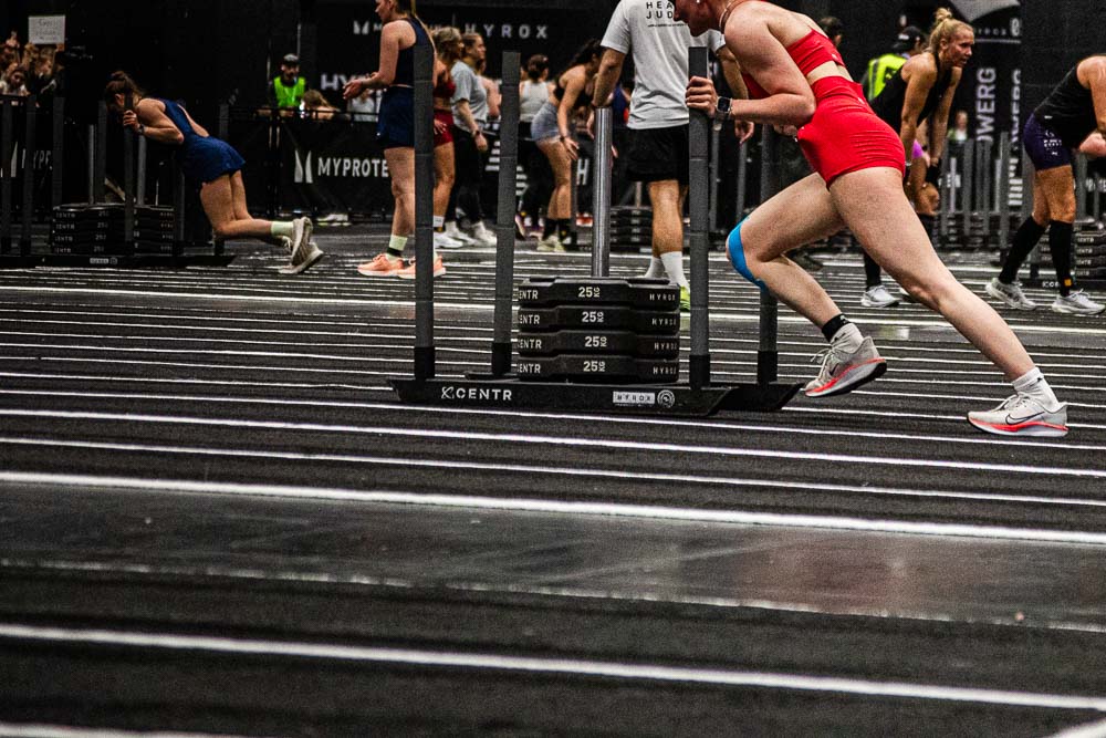 Atlete in een rode sportoutfit duwt tijdens een HYROX-wedstrijd een zware trainingsslee over het gemarkeerde indoorvlak, terwijl andere deelnemers op de achtergrond aan hun stations werken