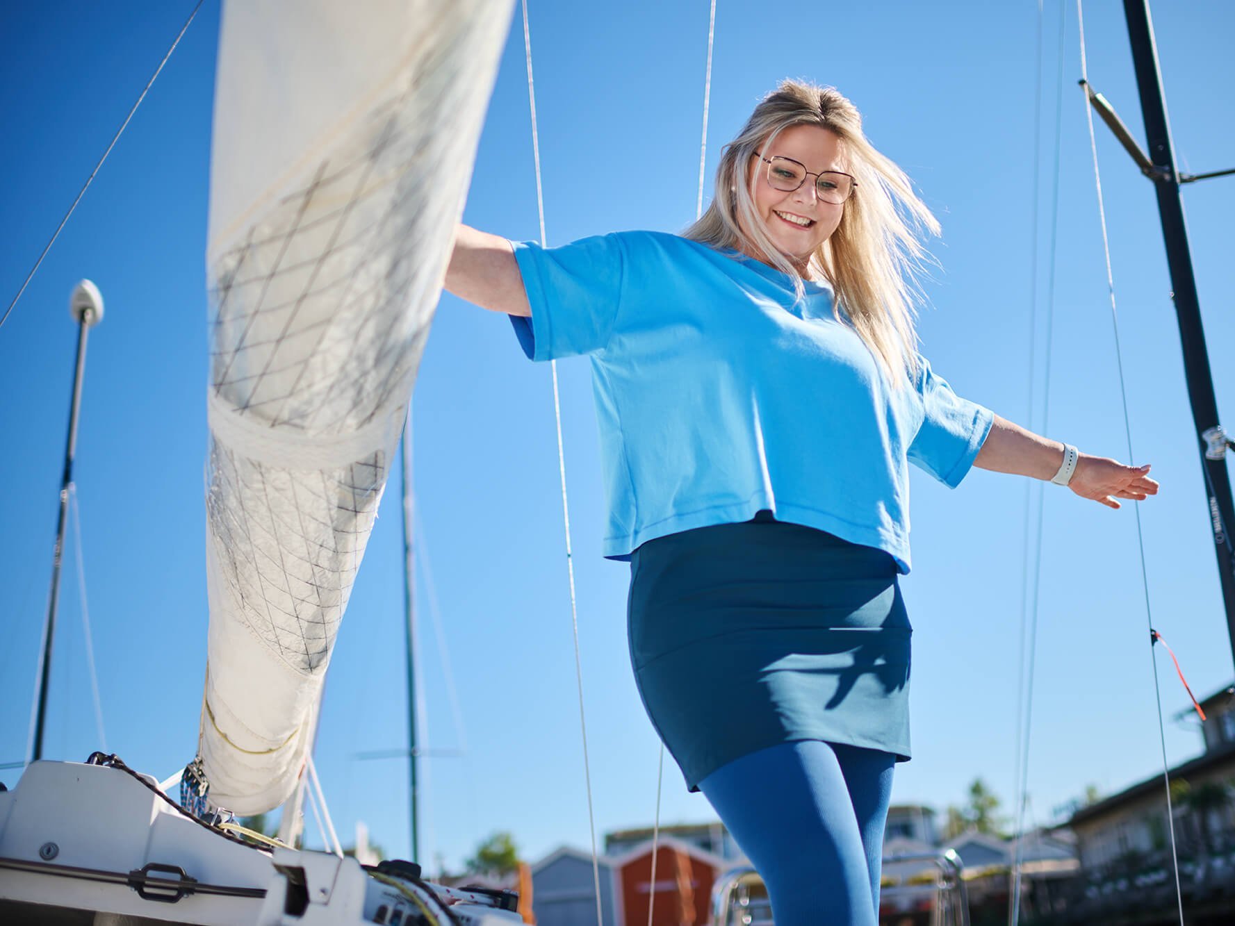 Vrouw in een blauw T-shirt en donkere rok met de curaflow lip- en lymfeverzorging van Bauerfeind staat met gespreide armen op een zeilboot onder een heldere hemel.