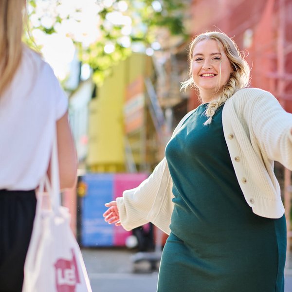 Twee vrouwen komen elkaar tegen op een zonnige straat in een stad. Een vrouw met een blonde vlecht, groene jurk en wit vest spreidt vrolijk haar armen en lacht naar de andere vrouw. Op de achtergrond zijn kleurrijke steigers en gebouwen te zien.