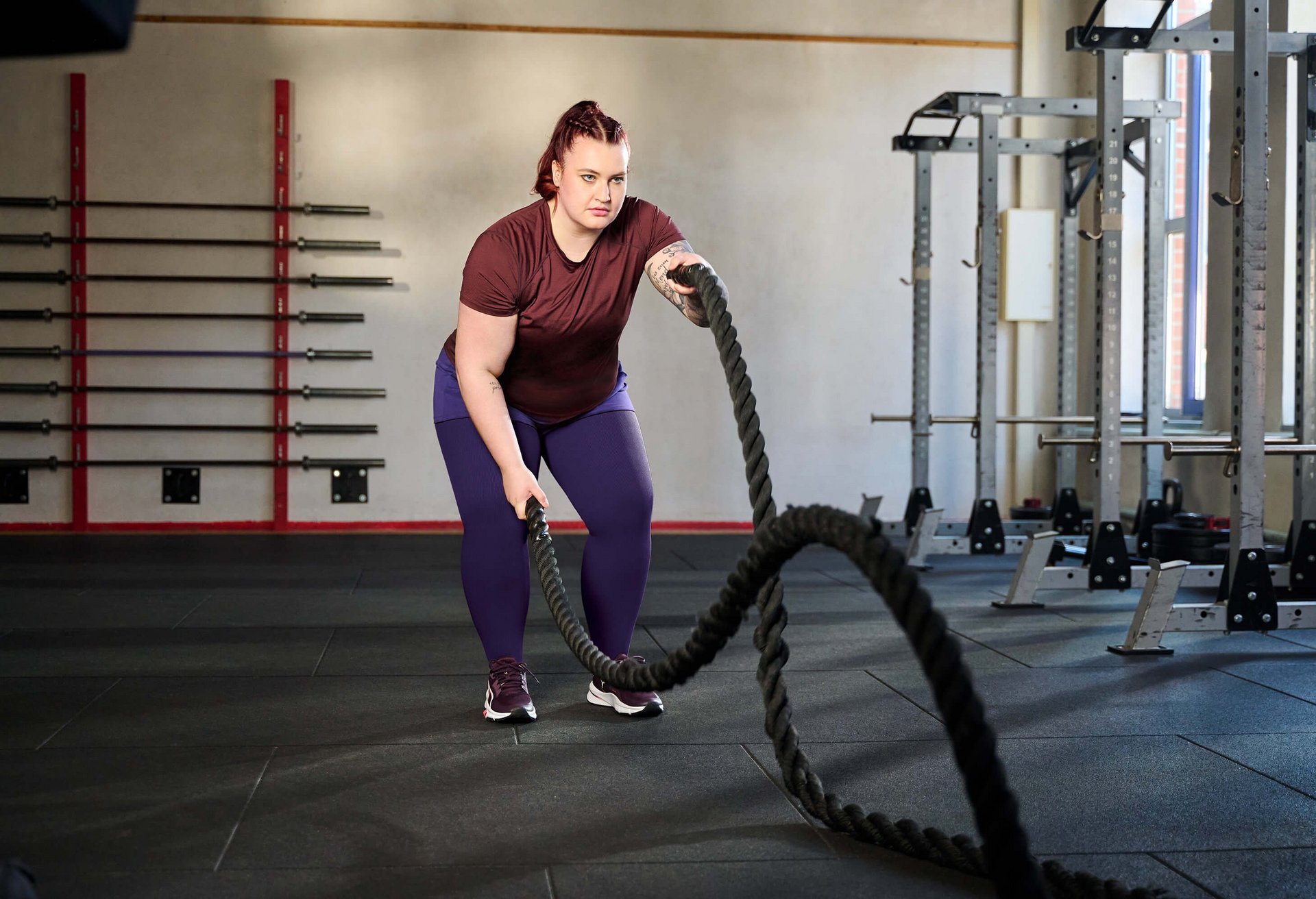 Een vrouw traint in een fitnessstudio met battle ropes. Ze draagt een bordeauxrood T-shirt, een paarse legging en sportschoenen. Op de achtergrond zijn halters aan de wand en krachttrainingsapparaten te zien.