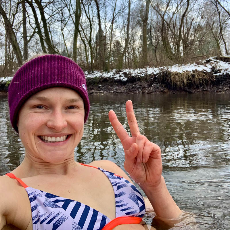 Anna Hahner staat tot haar borst in het koude water, omringd door een besneeuwd landschap en kale bomen, glimlachend en een peace-gebaar makend met haar vingers.