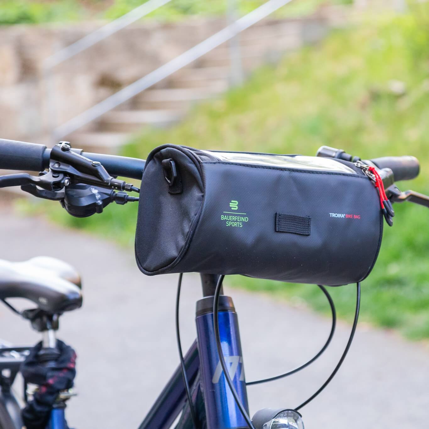 Zwarte Bauerfeind Sports TROIKA Bike Bag bevestigd aan het stuur van een blauwe fiets, met groen Bauerfeind Sports logo, transparant smartphonevak aan de bovenkant en rode ritstrekkers – praktisch georganiseerd voor ontspannen ritten en actieve avonturen.