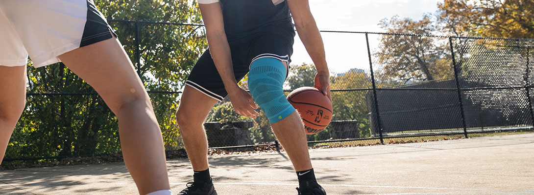 Een basketbalspeler staat op een betonnen veld dat is omgeven door een hek en doet een truc met de bal door hem door zijn twee benen te laten gaan. Een andere speler staat tegenover hem. Hij draagt een kniekous.