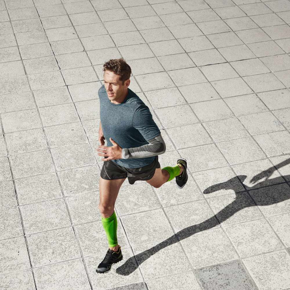 Een fitte hardloper in een blauw shirt en zwarte shorts sprint over een bestrate ondergrond. Hij draagt groene compressiekousen voor betere doorbloeding en spierondersteuning. Zijn houding straalt energie en dynamiek uit.