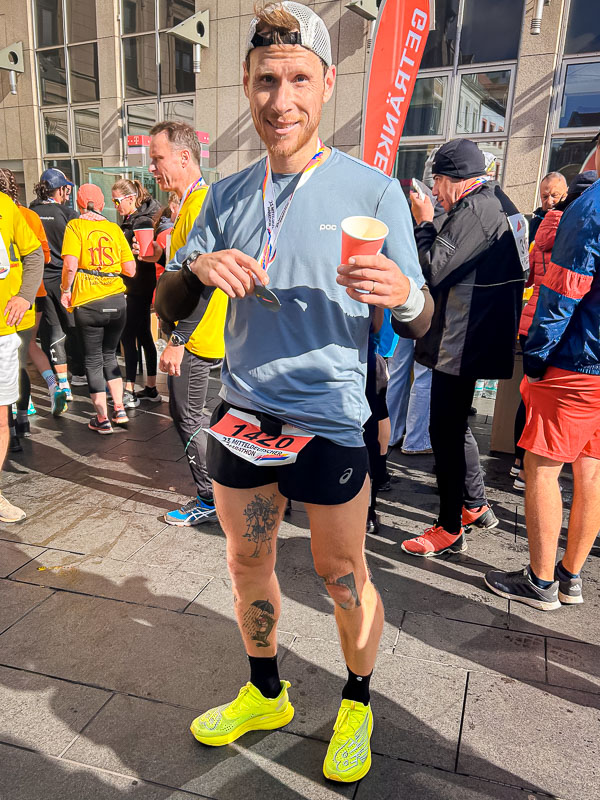 Trots bij de finish na het voltooien van de halve marathon in Halle, met een medaille en een drankje in de hand, genietend van het moment na de race.