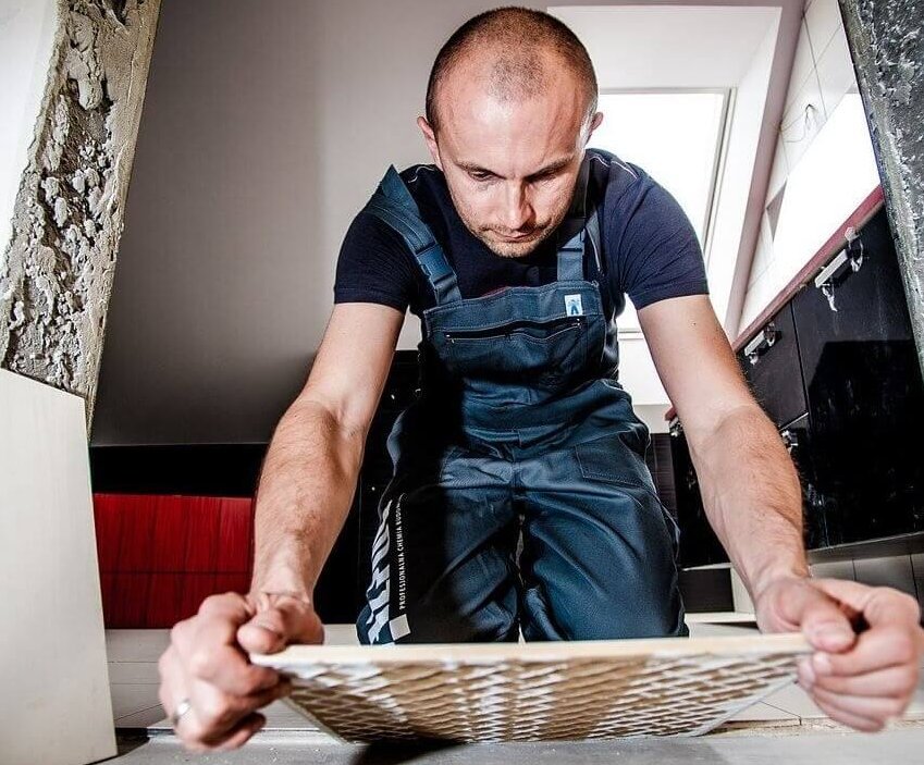 De afbeelding toont een klusser in knielende houding tijdens het tegelleggen. De hoge belasting voor de knieën tijdens het werk brengt het risico op gonartrose met zich mee.