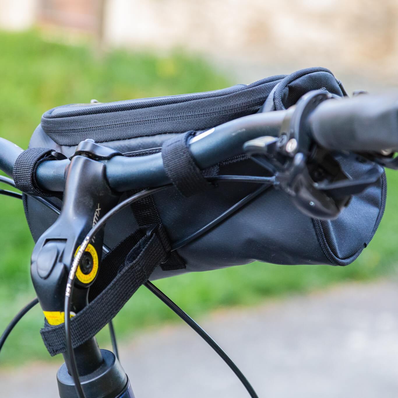 Close-up van de zwarte fietstas aan het stuur, waarbij bevestigingsriemen, remhendel, kabels en de bovenste ritssluiting goed zichtbaar zijn – de tas zit compact op het cockpitgedeelte en houdt belangrijke spullen onderweg binnen handbereik.