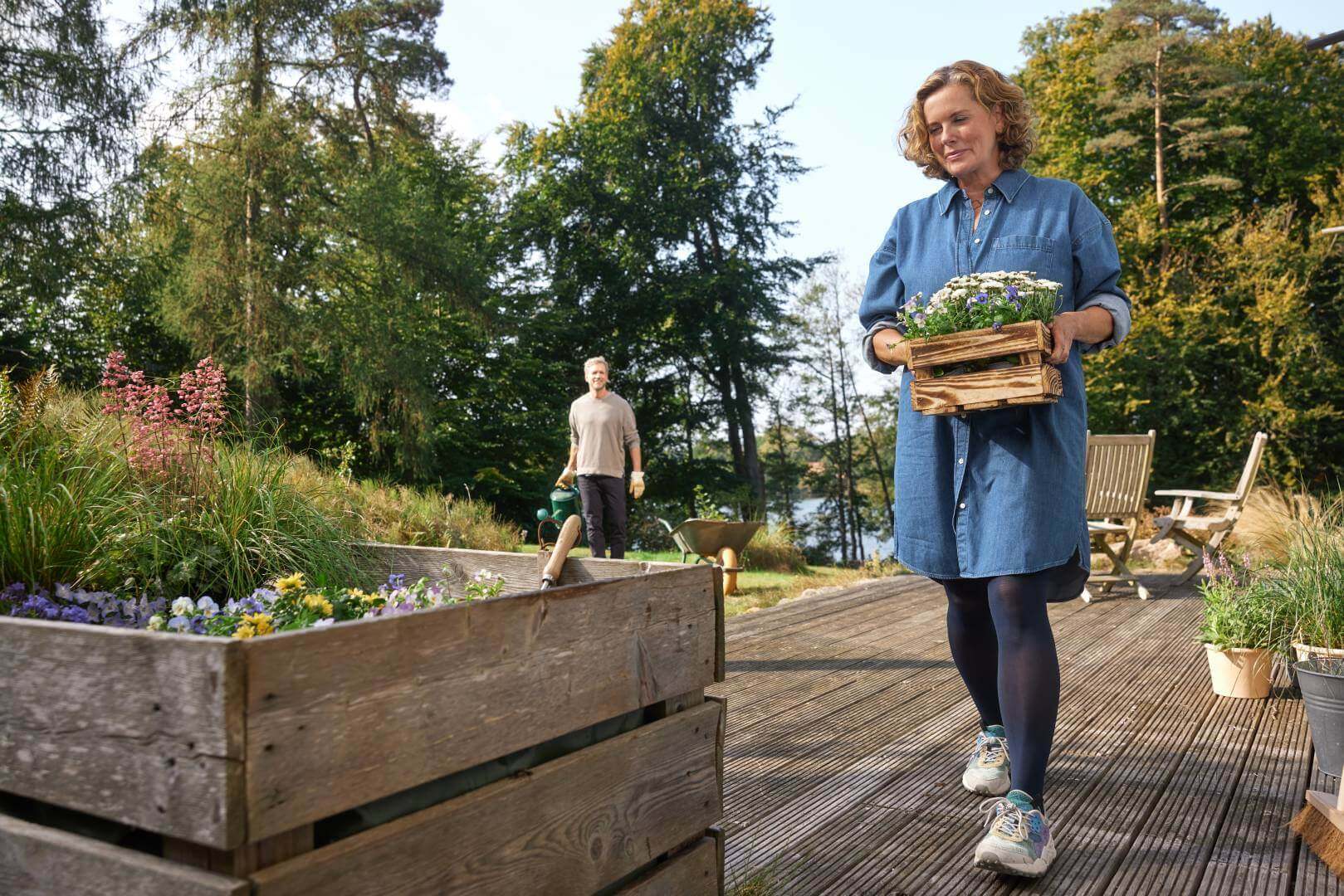 Een vrouw draagt een bloembak over een houten terras, terwijl een man op de achtergrond met een gieter staat. De vrouw draagt VenoTrain soft compressiekousen en een spijkerjurk met sportschoenen.
