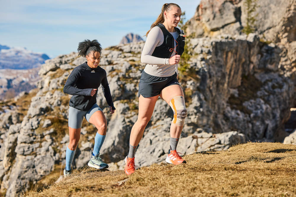 Twee sportieve vrouwen lopen bergopwaarts over een smal bergpad. De voorste loopster draagt een lichte outdoor compressie-kniebandage, terwijl de tweede atlete vlak achter haar volgt. Het rotsachtige alpiene landschap vormt de achtergrond.