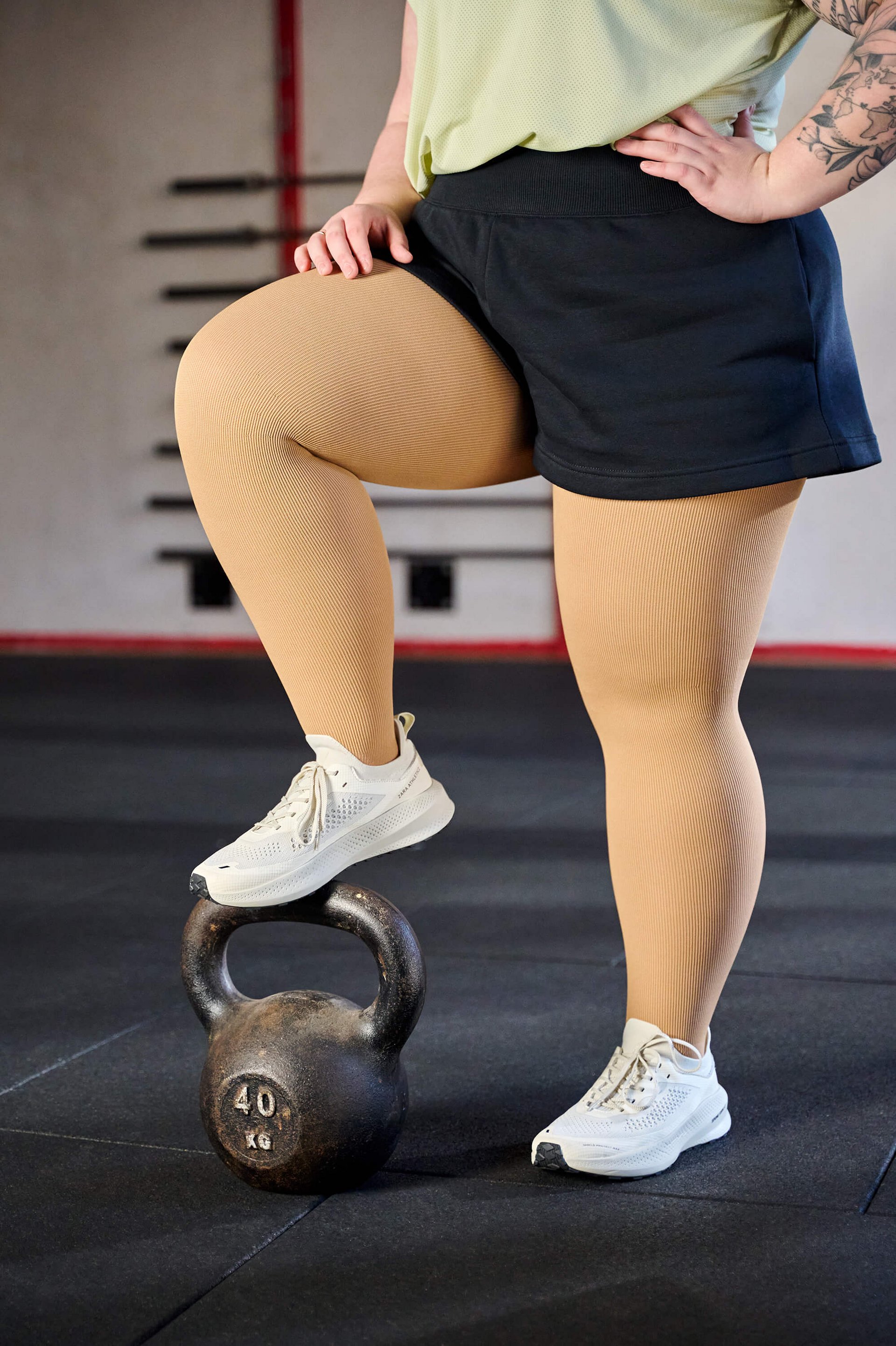 Close-up van de benen van een persoon in crèmekleurige compressiekousen en witte sportschoenen. De persoon draagt een zwarte short en heeft één been op een kettlebell van 40 kg in een fitnessstudio gezet.