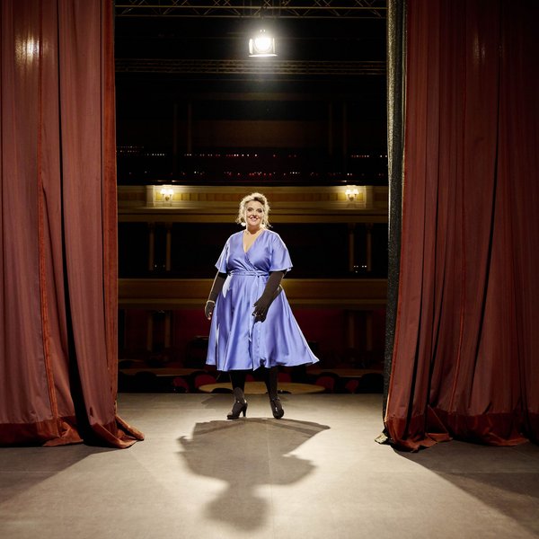 Een vrouw staat in een elegante, blauwe jurk op een theaterpodium tussen twee rode gordijnen, frontaal beschenen door een schijnwerper. De theaterzaal op de achtergrond is zwak verlicht.