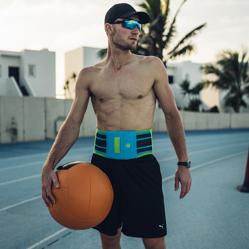 Een sportieve man met een pet en zonnebril staat op een atletiekbaan en draagt een blauwe rugbrace met groene accenten. Hij houdt een grote medicijnbal in zijn hand.