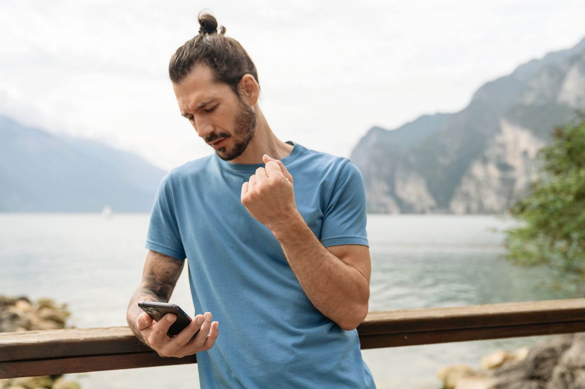 De afbeelding toont een man met smartphone die aan de oever van een bergmeer staat. Hij houdt zijn rechterarm met pijnlijke pols omhoog.