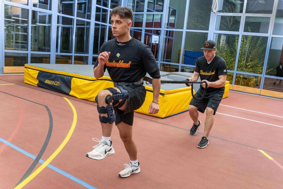 Honkballer Simon Liedtke doet weerstandstraining in een hal. Hij draagt de SecuTec Genu Flex knieorthese om zijn rechterbeen na zijn gescheurde achterste kruisband; achter hem houdt een trainer een weerstandstouw vast.